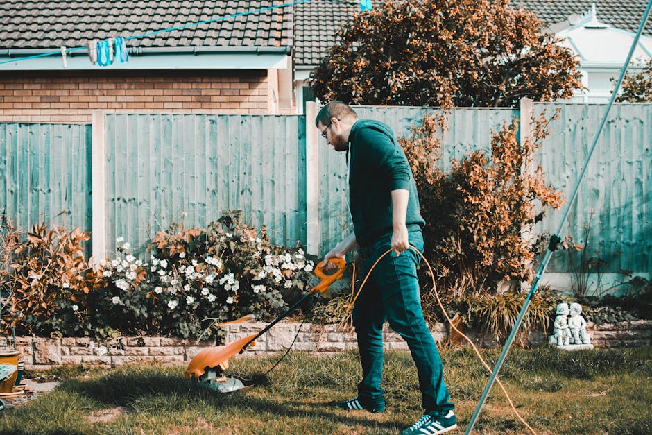 A man wearing glasses, a dark hoodie, and jeans is trimming grass with an orange electric lawn mower in a well-maintained garden. The garden features a neatly cut lawn bordered by flower beds filled w