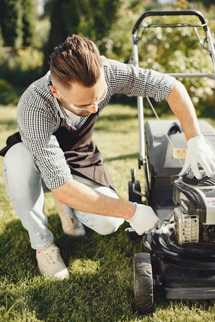 A man dressed in a checkered shirt, wearing safety goggles and white gloves, is kneeling on a well-maintained lawn while inspecting and performing maintenance on a petrol-powered lawn mower. The garde