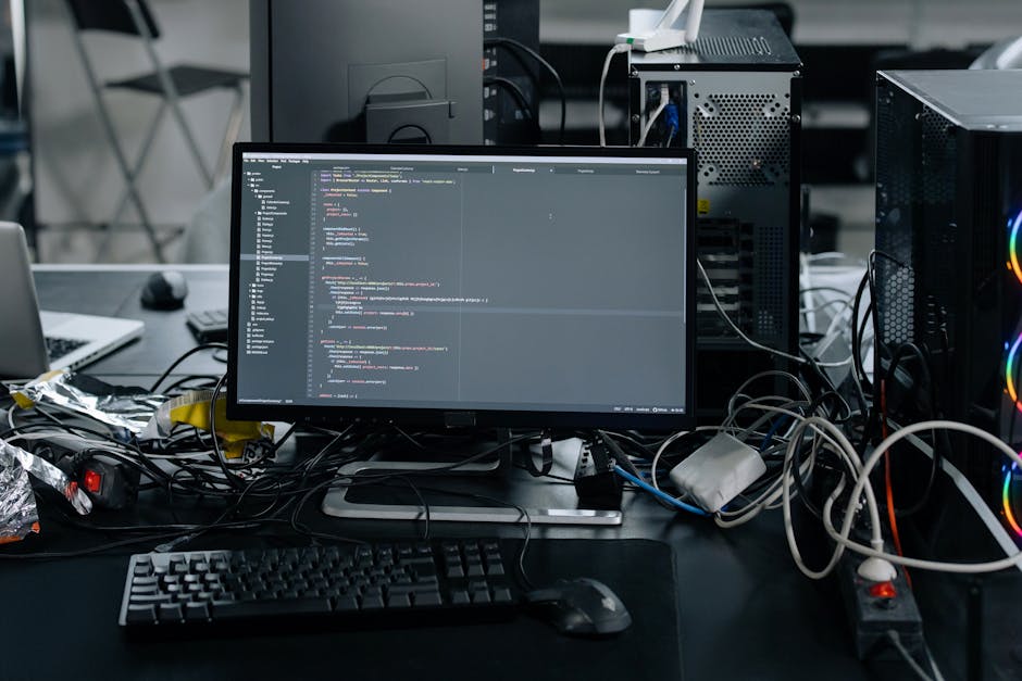 A cluttered workspace featuring a computer monitor displaying lines of code, surrounded by tangled wires, a keyboard, and a mouse on a dark desk. In the background, there are desktop towers with visib